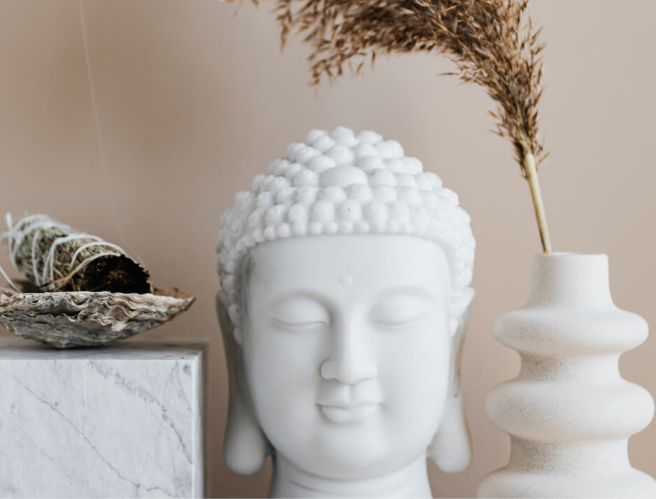A képen egy fehér Buddha-szobor látható, amely nyugodt és békés arckifejezést sugall. A kompozícióban mellette egy dekoratív váza szárított növénnyel, valamint egy márványhatású felületre helyezett füstölő vagy zsályaköteg található egy kagylóhéjban. Az összkép minimalista és harmonikus, a természetes színek és anyagok pedig a nyugalom és a meditáció érzését keltik.