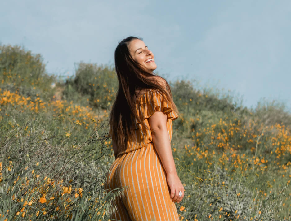 A képen egy fiatal nő látható egy napsütötte mezőn, tele sárga vadvirágokkal. Vidám, gondtalan mosollyal néz hátra, miközben a szél lágyan fújja hosszú haját. A sárga csíkos ruha harmonizál a természet színeivel, fokozva a melegség és szabadság érzetét. Az ég kékje és a virágok vibráló színei idilli, nyugodt hangulatot teremtenek.